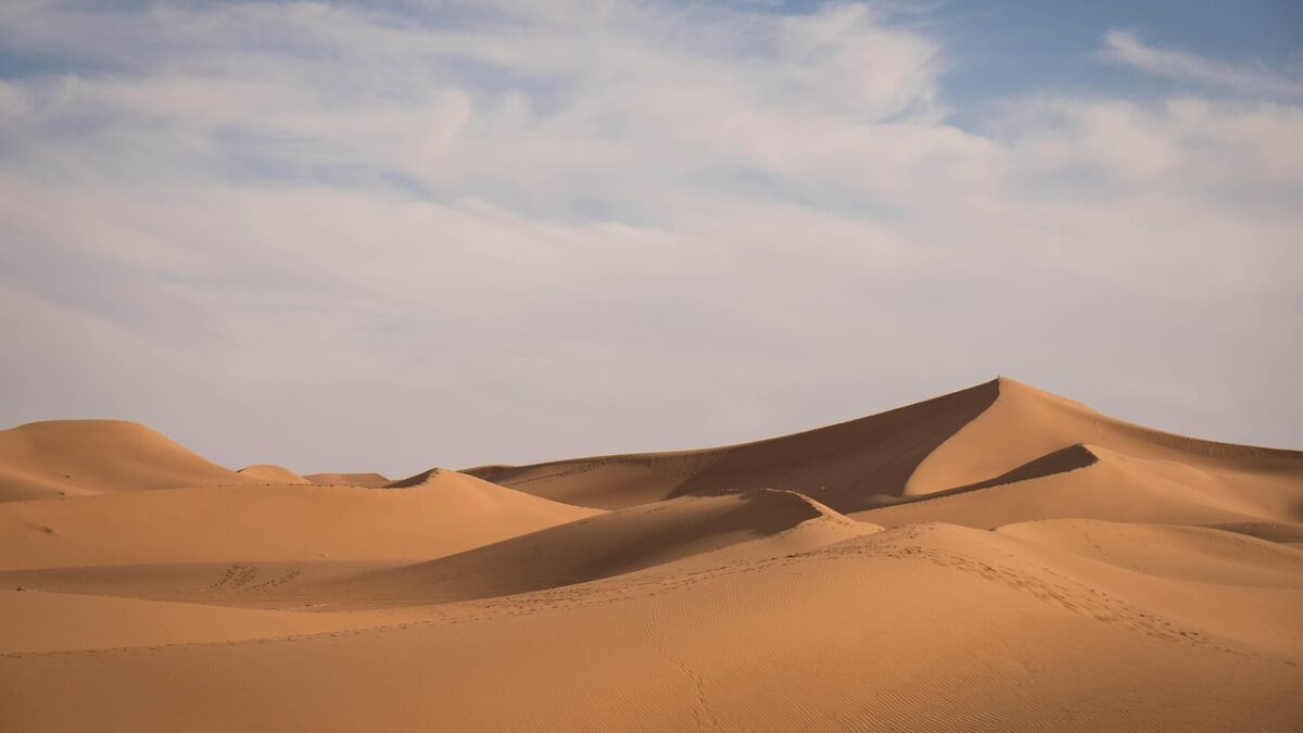 Dunes de sable orangé ondulantes sous un ciel bleu voilé de nuages, au relief doux et sans aucune présence humaine.