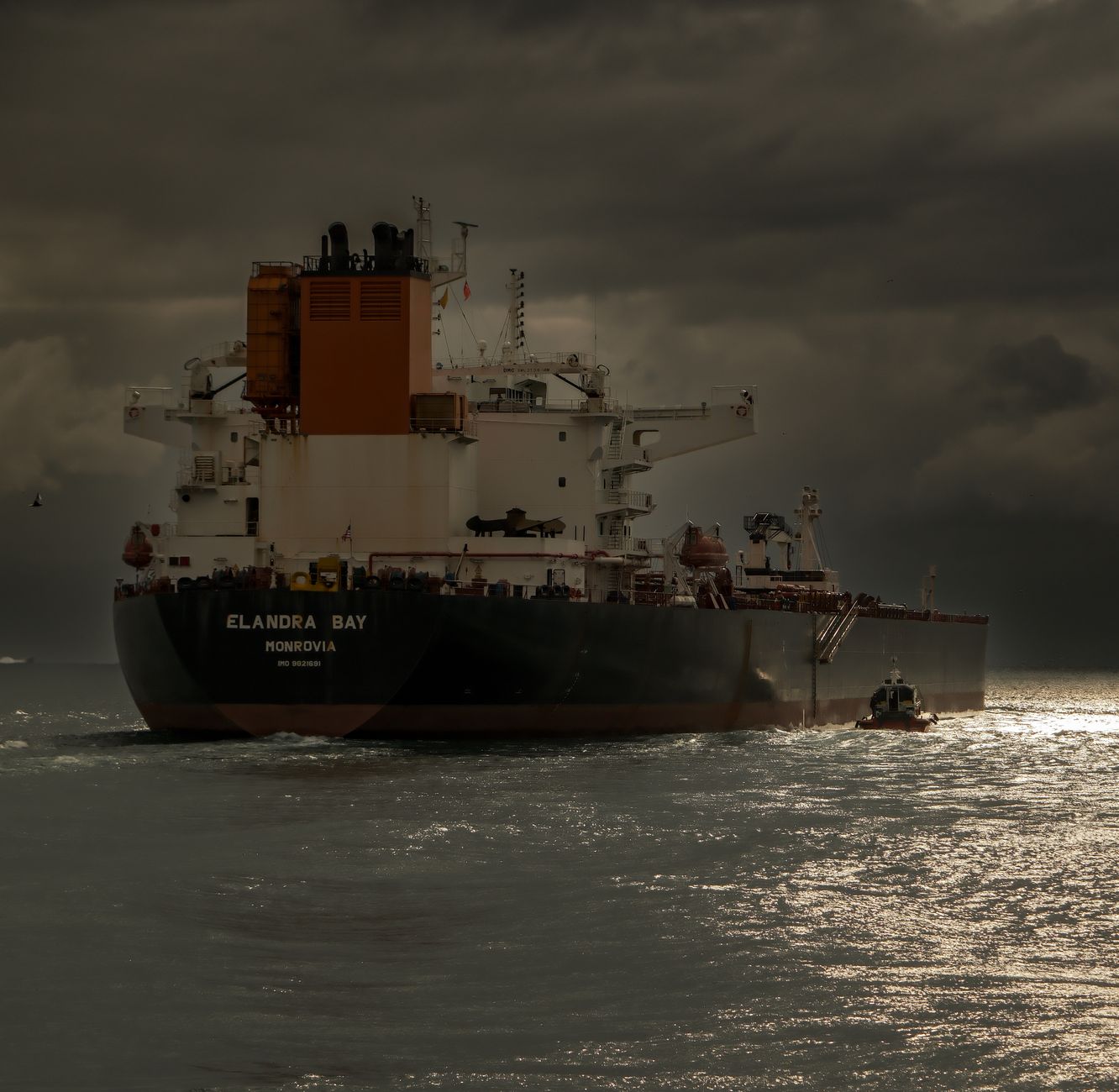 Grand navire-citerne vu de l’arrière en mer sous un ciel sombre et nuageux, avec un petit bateau près de la coque.